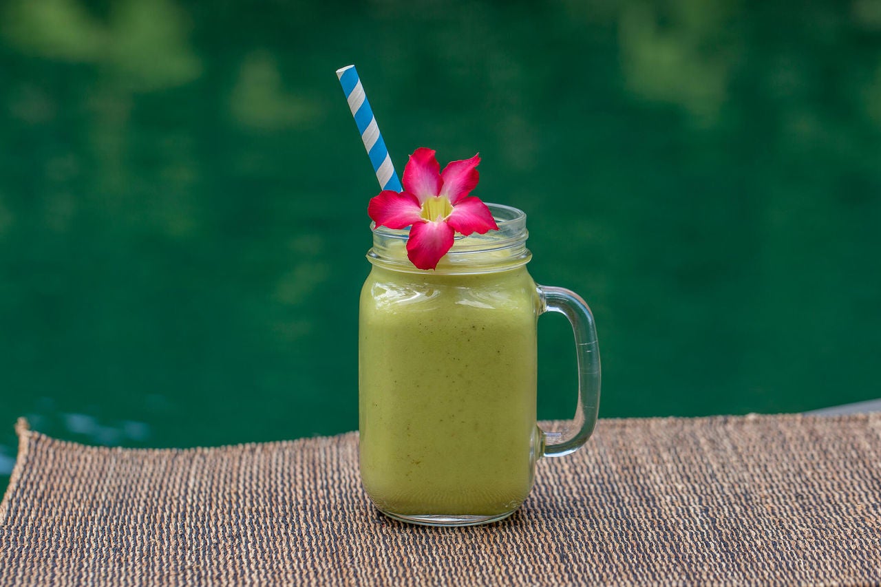 Tropical green smoothie with pink flower garnish and straw by water. 