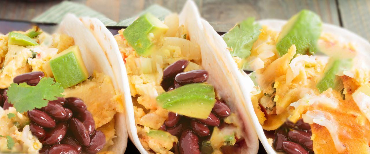 Three soft tacos filled with black beans, avocado chunks, vegetables, and fresh cilantro, served on a rustic wooden table.