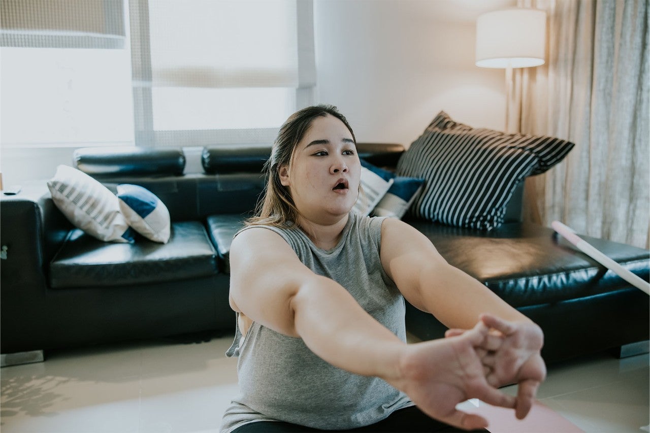 Focused woman stretching her arms while exercising at home, sitting on the floor in a living room with a black leather couch and decorative pillows.