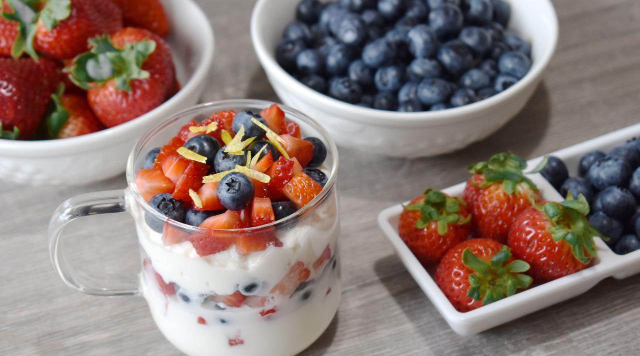 A glass cup filled with yogurt, blueberries, and strawberries sits on a table next to bowls of fresh strawberries and blueberries.