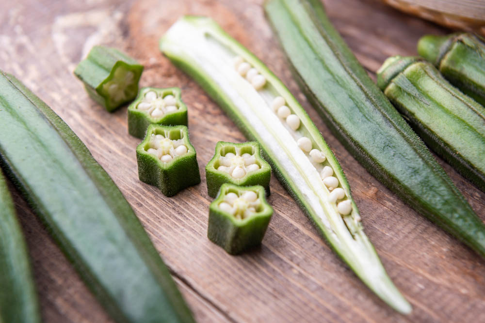 Fresh okra pods are displayed on a wooden surface, with some sliced open to reveal their seeds.