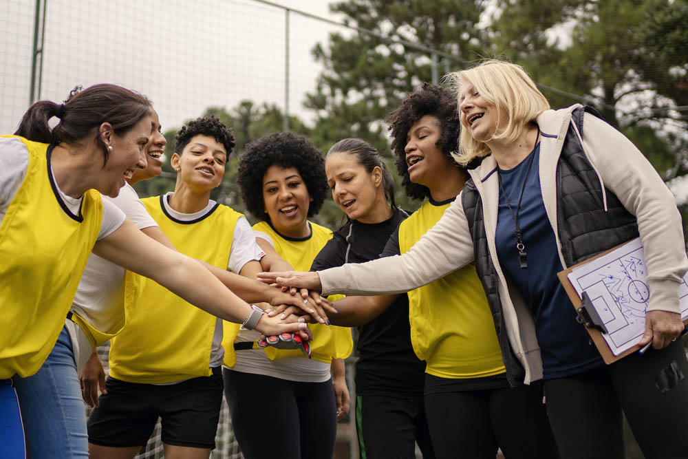 A group of women wearing yellow jerseys and a coach are gathered on a sports field, smiling and putting their hands together in a gesture of team spirit and camaraderie.