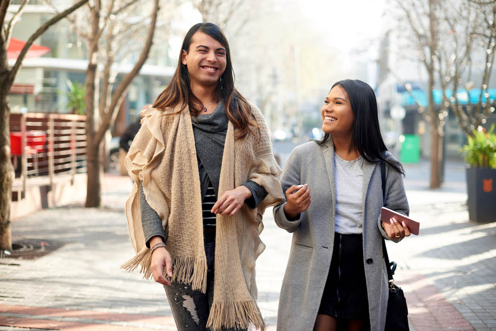 Two individuals are walking side by side down a city street, smiling and enjoying each other's company, surrounded by trees and urban scenery.