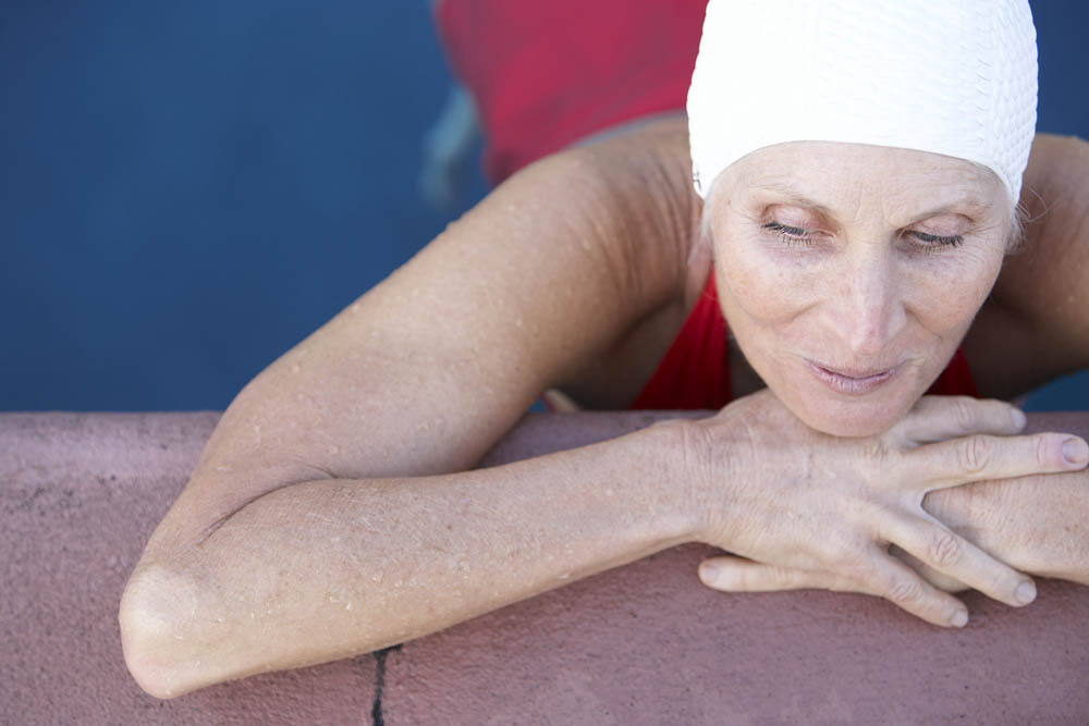 Elderly woman in a red swimsuit and white swim cap resting her arms on the edge of a swimming pool, looking down with a peaceful expression, with blue water in the background.