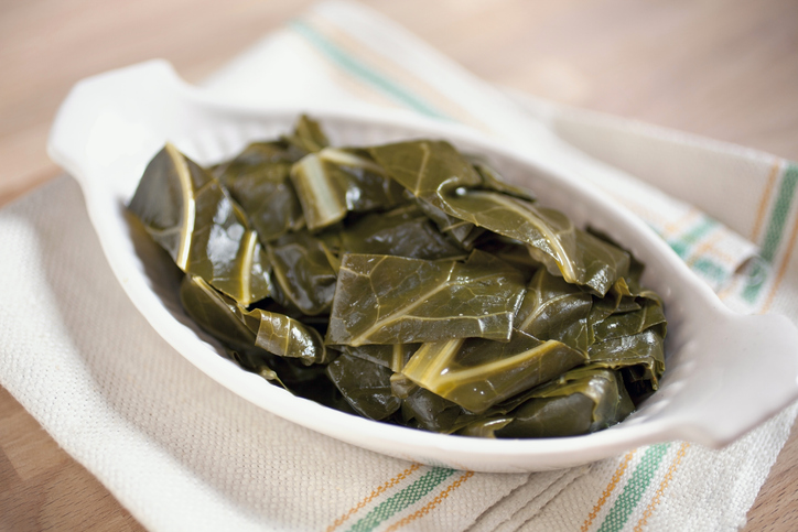 White ceramic dish filled with tender, cooked collard greens, served on a striped kitchen towel on a wooden surface.
