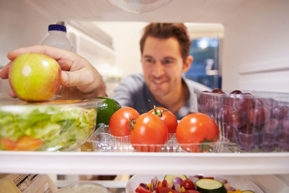A man is reaching into a refrigerator, picking up a green apple while surrounded by various fresh foods, including tomatoes, salad, and grapes, with a cheerful expression.