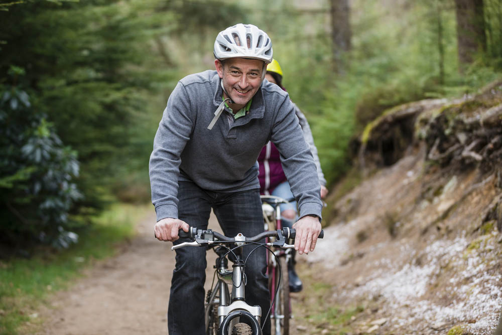Smiling man wearing a helmet, riding a bicycle on a forest trail.