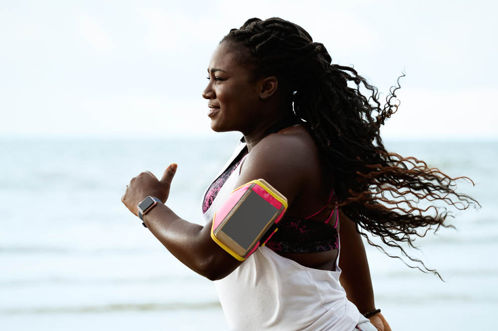 A woman with long, flowing hair runs along a beach, wearing a fitness armband for her smartphone and an athletic outfit, with the ocean in the background.