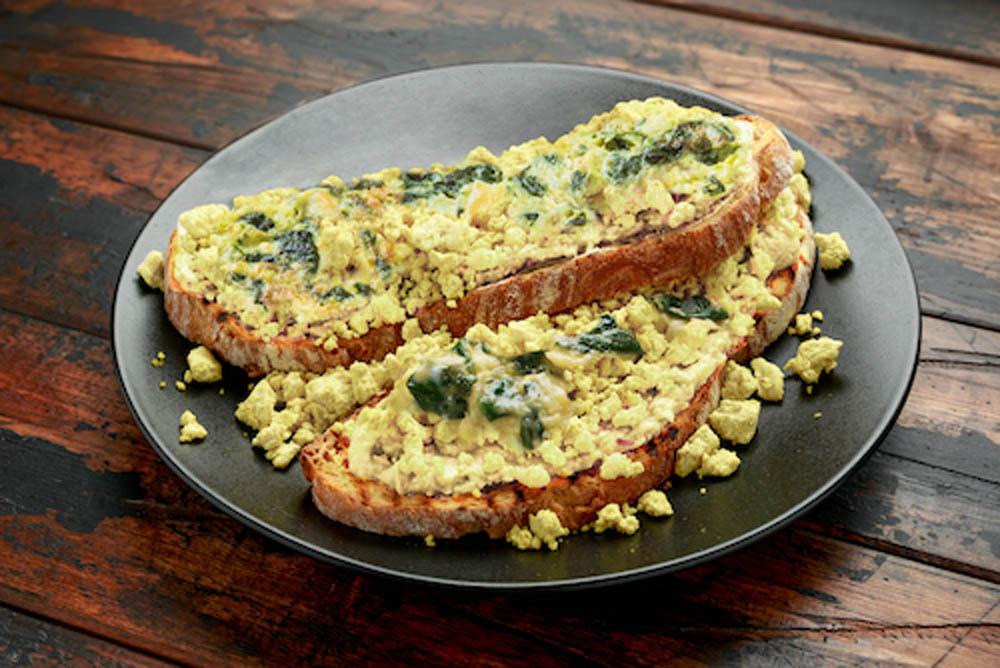 Scrambled tofu with spinach served on two slices of toasted bread, placed on a dark plate on a rustic wooden table