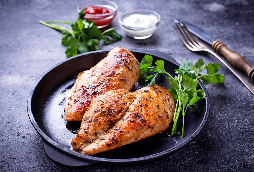 Two grilled chicken breasts with a golden-brown, herb-seasoned crust, served in a black cast iron skillet, garnished with fresh parsley, with forks and dipping sauces in the background.