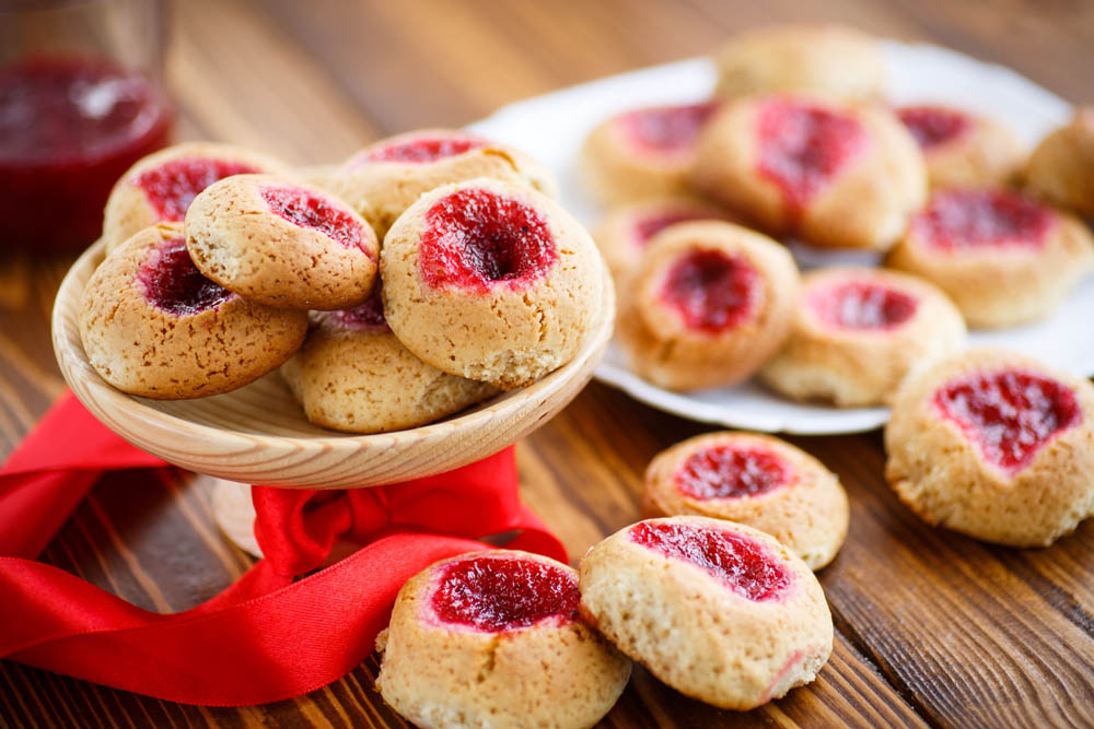 A wooden bowl filled with thumbprint cookies topped with red jam, surrounded by more cookies on a plate and a glass of jam in the background, all set on a wooden table with a red ribbon.