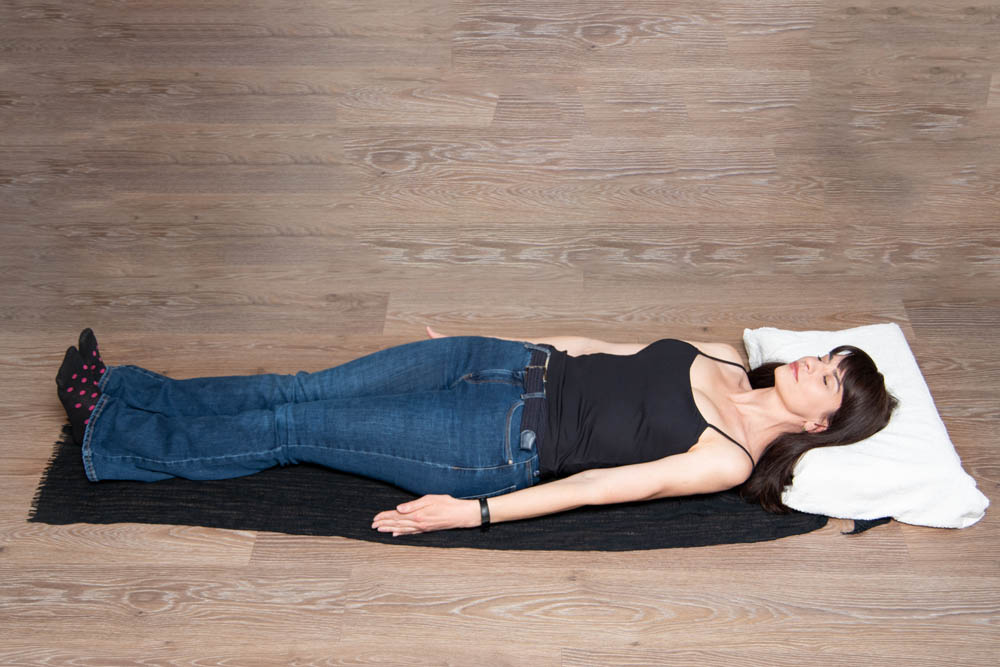 A woman lying on her back on a black yoga mat, dressed in a black tank top and blue jeans, with her arms extended and a pillow under her head, in a calm and relaxed position on a wooden floor.
