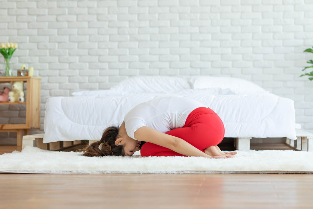 A woman in a yoga pose, resting her forehead on her knees while sitting on a soft rug in a bright room with a bed in the background, creating a peaceful atmosphere.