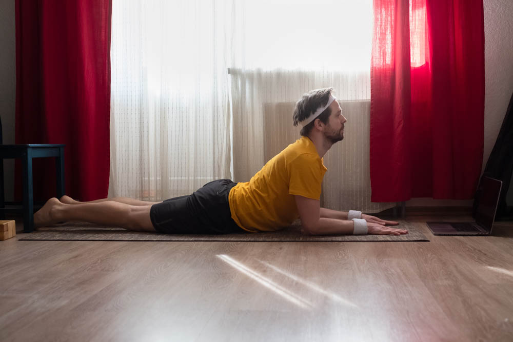 A man performing a yoga pose, lying on his stomach with his head lifted, wearing a yellow shirt and wristbands, in a room with red curtains and natural light streaming in.