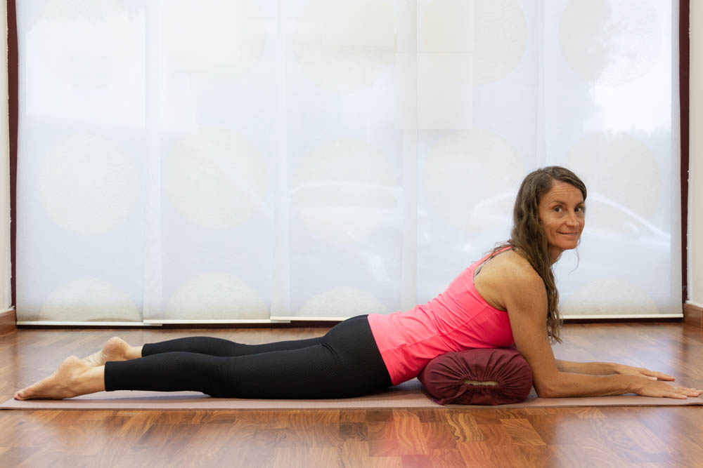 A woman lying on her stomach on a yoga mat, resting her forearms on a bolster and looking towards the camera, with light filtering through sheer curtains in the background.