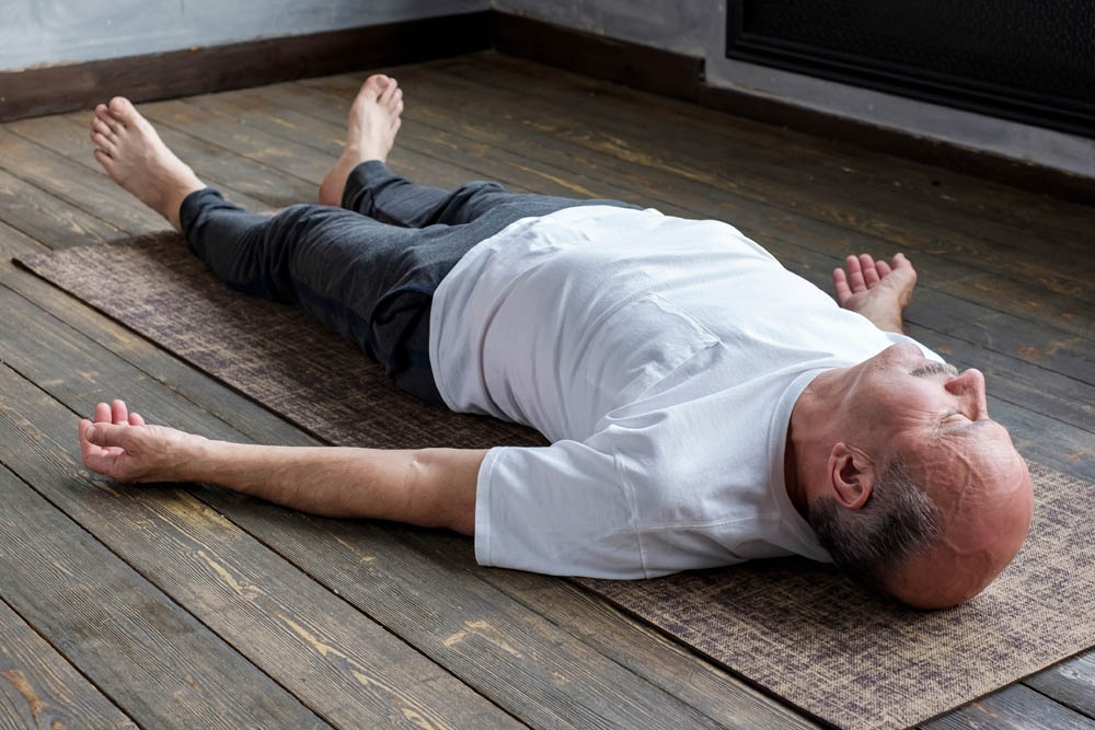 An elderly man lying flat on his back on a yoga mat, dressed in a white shirt and gray pants, with his arms extended out to the sides and a relaxed expression, on a wooden floor.
