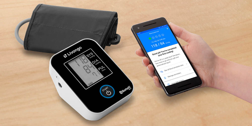 A blood pressure monitor sits on a wooden surface next to a smartphone displaying health data. A hand is holding the phone, indicating a connection between the devices.
