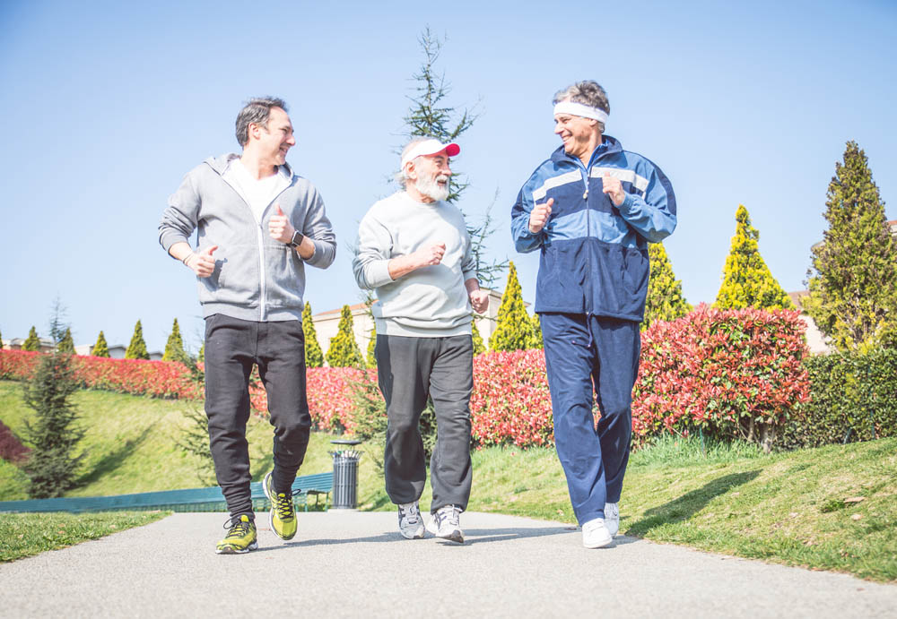 three men of varying ages are jogging together on a path in a park, smiling and enjoying each other's company, surrounded by colorful flowers and greenery.