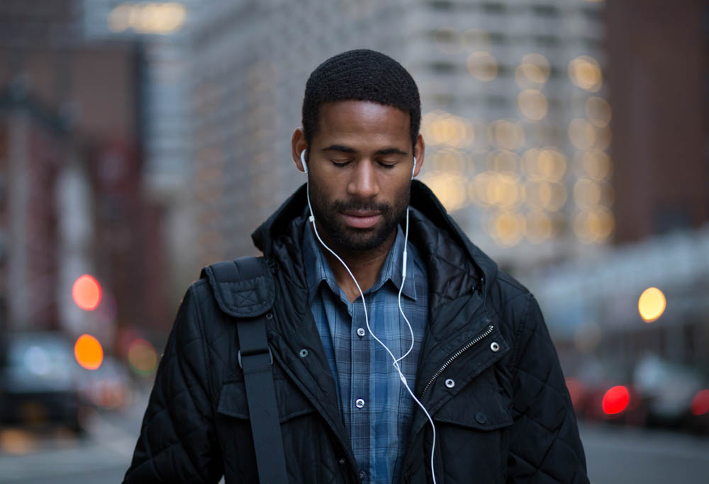 A man is walking through a city street, wearing headphones and looking down thoughtfully, with a blurred background of city lights and buildings.