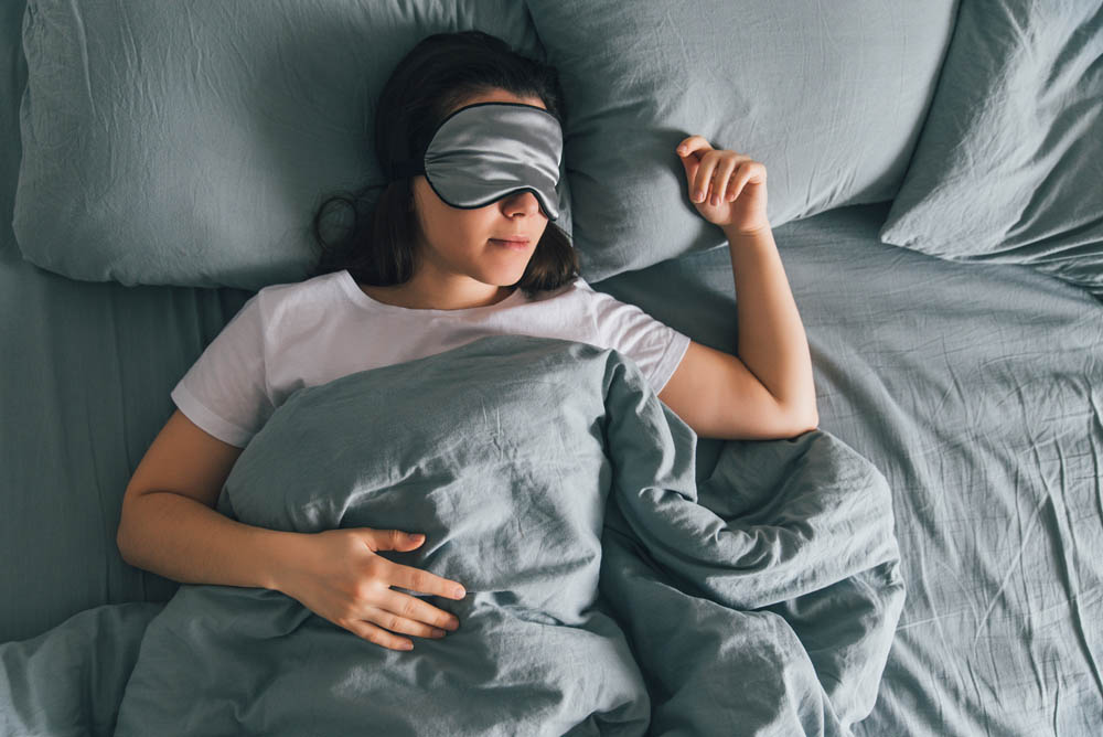 A woman wearing a sleep mask is resting in bed, surrounded by soft, blue bedding, appearing relaxed and peaceful.