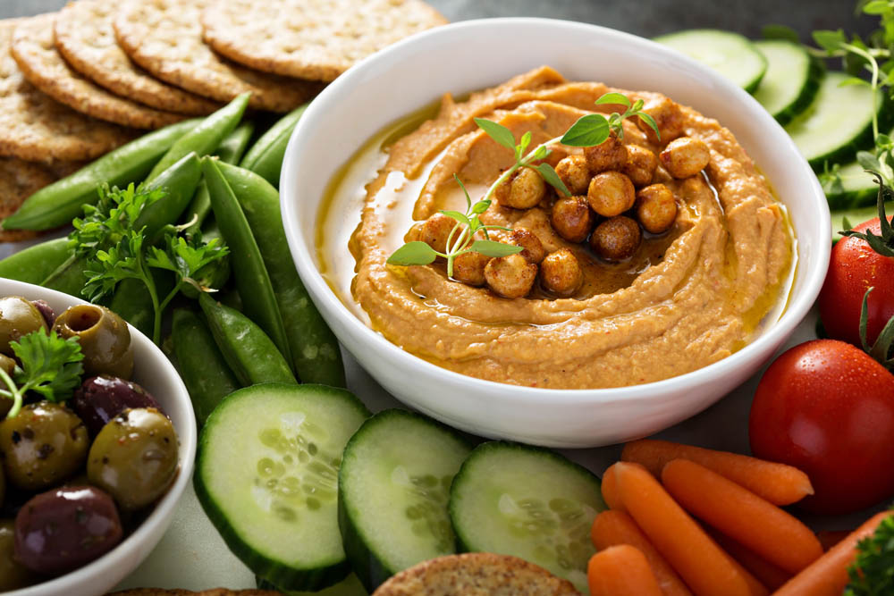 A vibrant spread of fresh vegetables and snacks, featuring a bowl of hummus topped with chickpeas and herbs, surrounded by cucumber slices, carrots, snap peas, olives, and crackers.