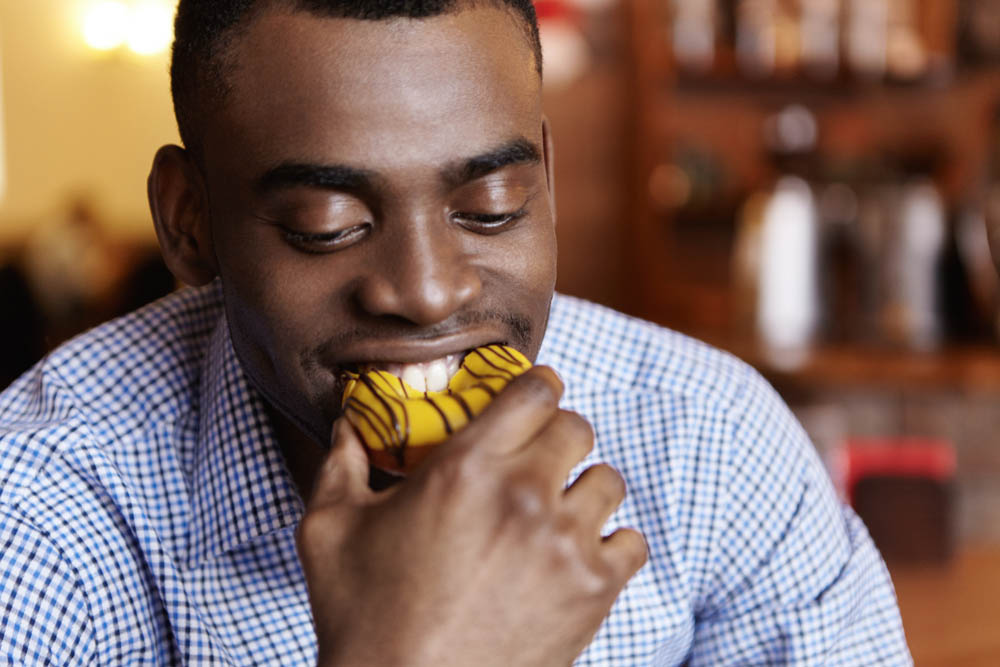 A smiling man is about to take a bite of a colorful dessert, holding it close to his mouth in a cozy café setting.