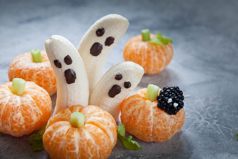 Halloween-themed fruit snack featuring banana ghosts with chocolate faces and peeled mandarin pumpkins with celery stems, arranged on a textured surface.