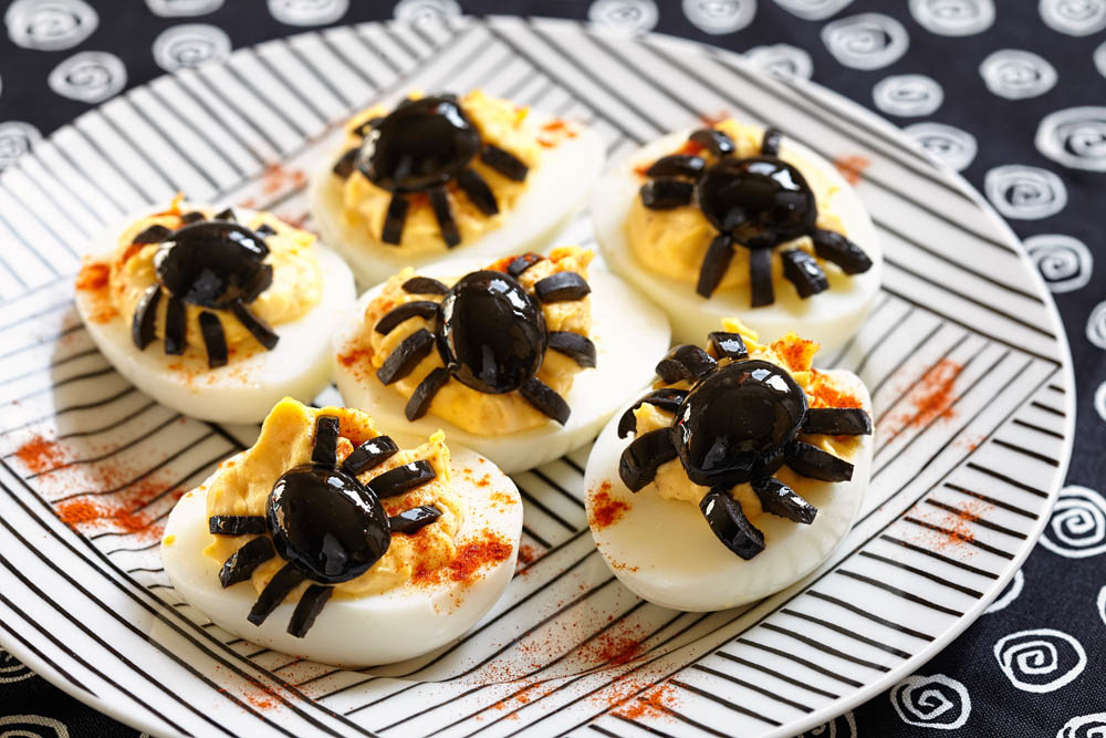 Deviled eggs decorated with black olive spiders, placed on a plate with paprika seasoning for a spooky Halloween treat.