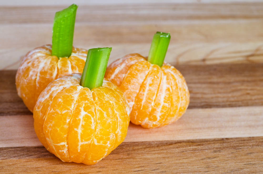Simple and healthy Halloween snack made of peeled mandarins with celery stems, resembling small pumpkins, placed on a wooden surface.