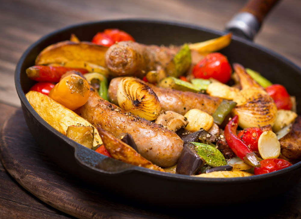 A cast iron skillet filled with roasted sausages, potatoes, and a variety of colorful vegetables, including cherry tomatoes, bell peppers, and onions, arranged artfully.