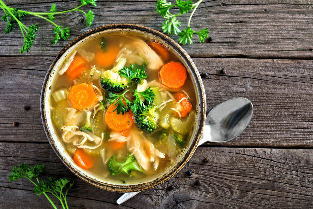 A bowl of chicken noodle soup filled with vegetables like carrots and broccoli, garnished with fresh parsley, resting on a wooden surface with a spoon beside it.