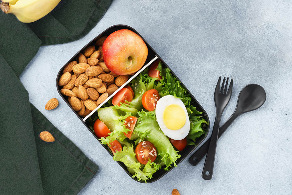 A black lunchbox divided into sections containing a salad with lettuce, cherry tomatoes, and a boiled egg, along with an apple and a handful of almonds, set on a gray surface with utensils nearby.