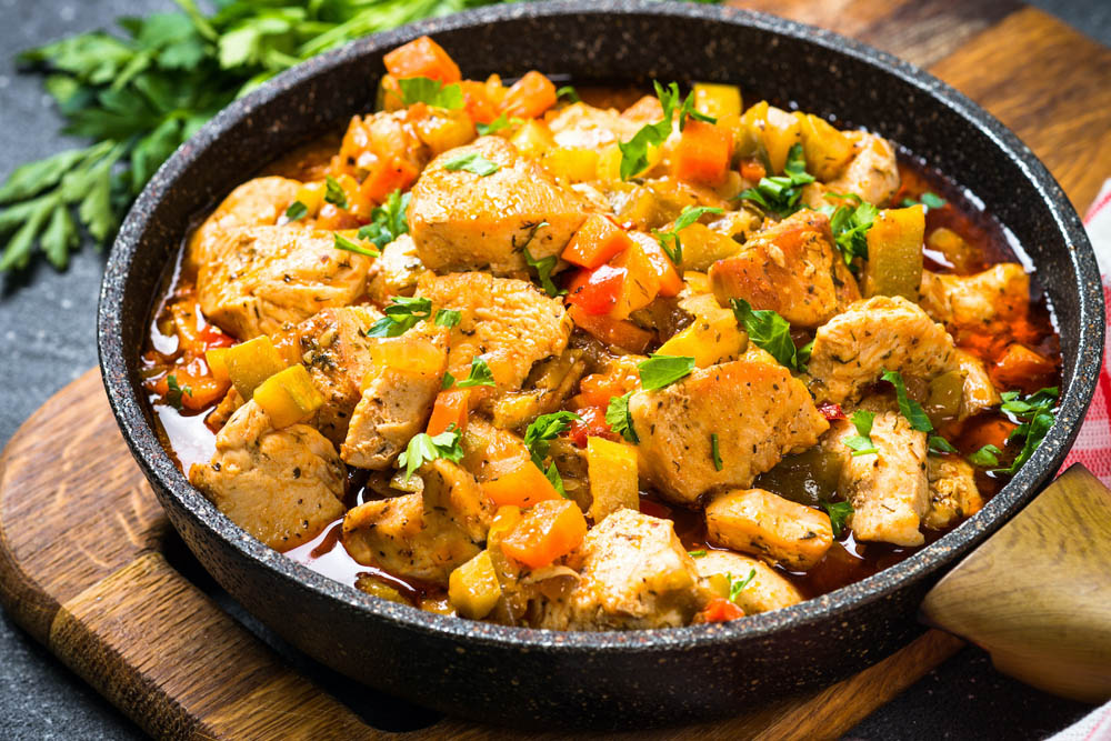 A cast iron skillet filled with sautéed chicken pieces mixed with diced tomatoes and bell peppers, garnished with fresh herbs, sitting on a wooden cutting board.