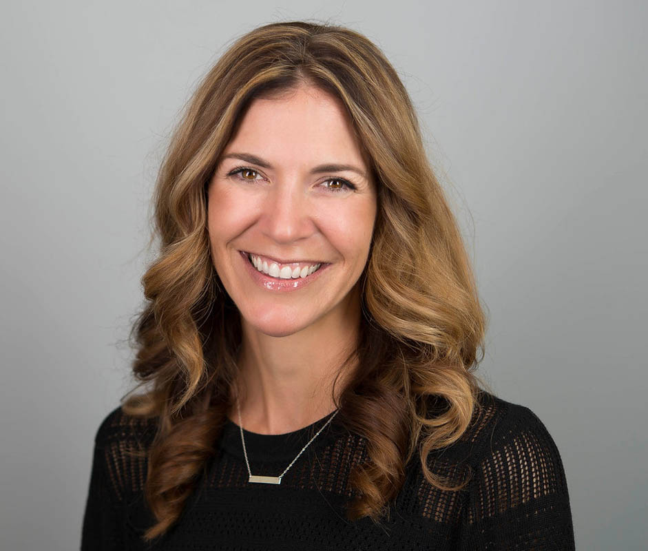 JA smiling woman with long, wavy hair poses for a professional headshot against a neutral gray background, wearing a black top and a simple necklace.