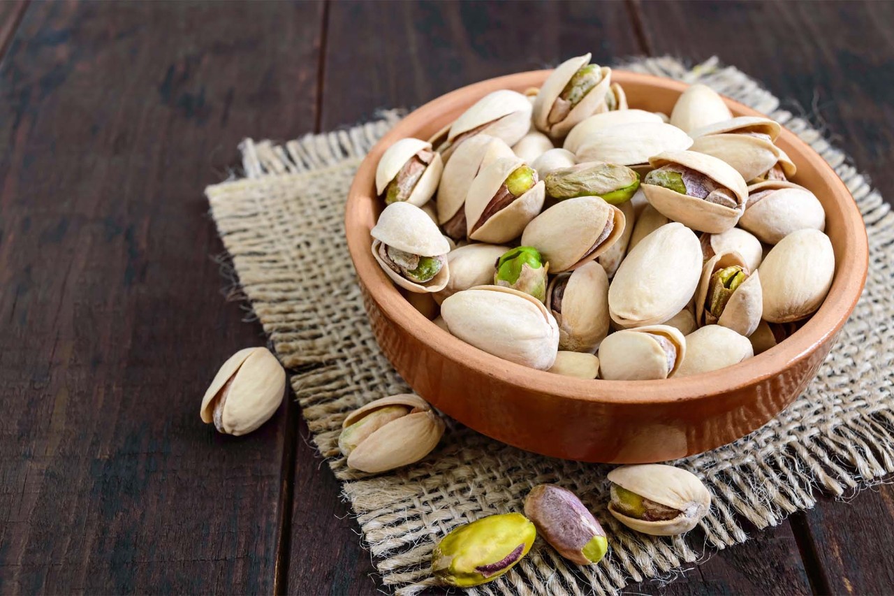 A wooden bowl filled with pistachios, some of which are open, placed on a burlap mat on a dark wooden surface, with a few pistachios scattered around.