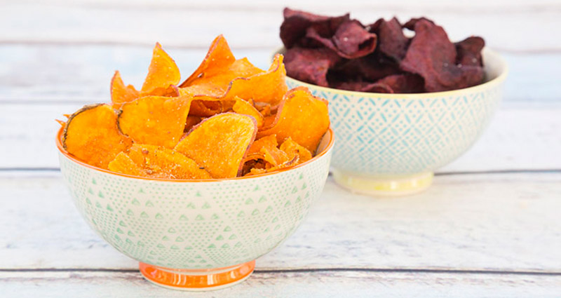 Two bowls on a wooden surface. The bowl on the left contains orange sweet potato chips, while the bowl on the right has purple beet chips.