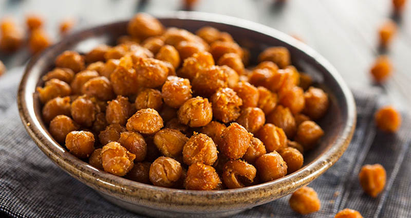 A bowl filled with roasted chickpeas on a textured surface. The chickpeas are golden brown and crispy, with some scattered around the bowl.