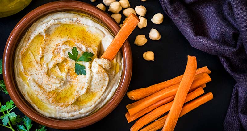 Bowl of hummus topped with olive oil and parsley, garnished with a carrot stick. Surrounded by chickpeas and more carrot sticks on a dark surface.
