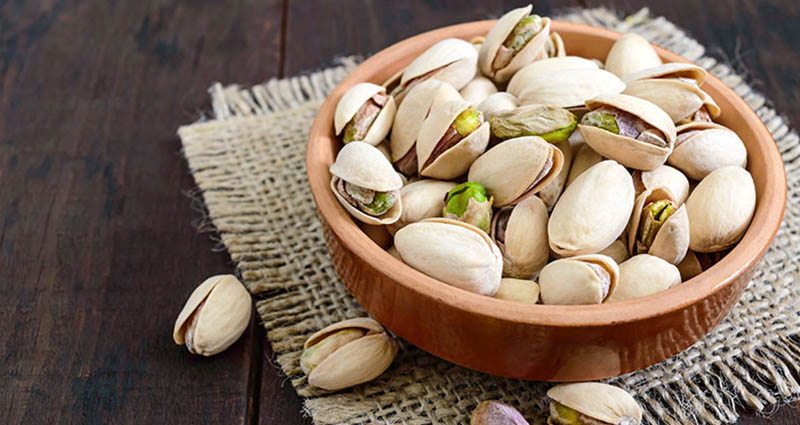 A wooden bowl filled with unshelled pistachios on a piece of burlap, set on a dark wooden surface.