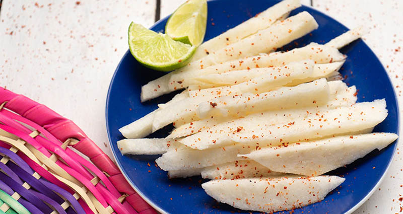 Sliced jicama on a blue plate, sprinkled with chili powder, accompanied by a lime wedge. A colorful woven mat is partially visible.