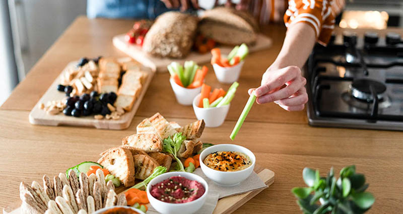 A person reaching for a celery stick from a platter of assorted snacks, including dips and fresh vegetables, arranged on a wooden table, with bread and olives in the background.