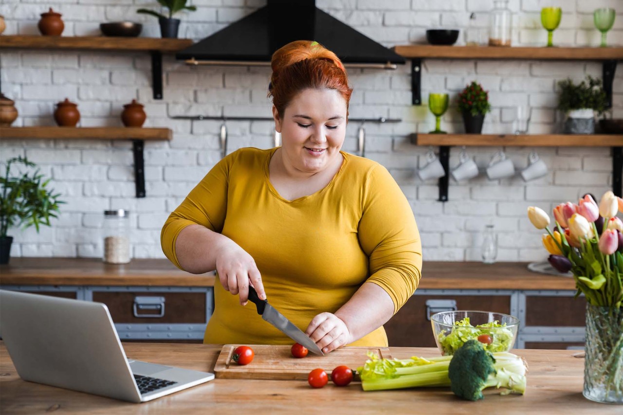 A woman in a yellow shirt chopping vegetables on a wooden kitchen counter, with a laptop open beside her and a vase of flowers in the foreground, set against a modern kitchen backdrop.