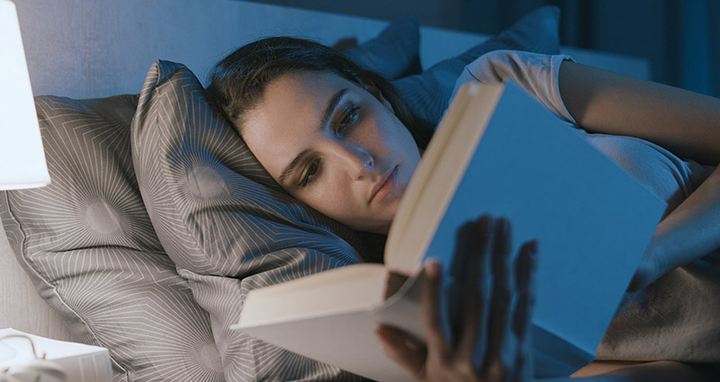 Young woman lying in bed, reading a book under soft bedside lamp light, with a calm and focused expression.