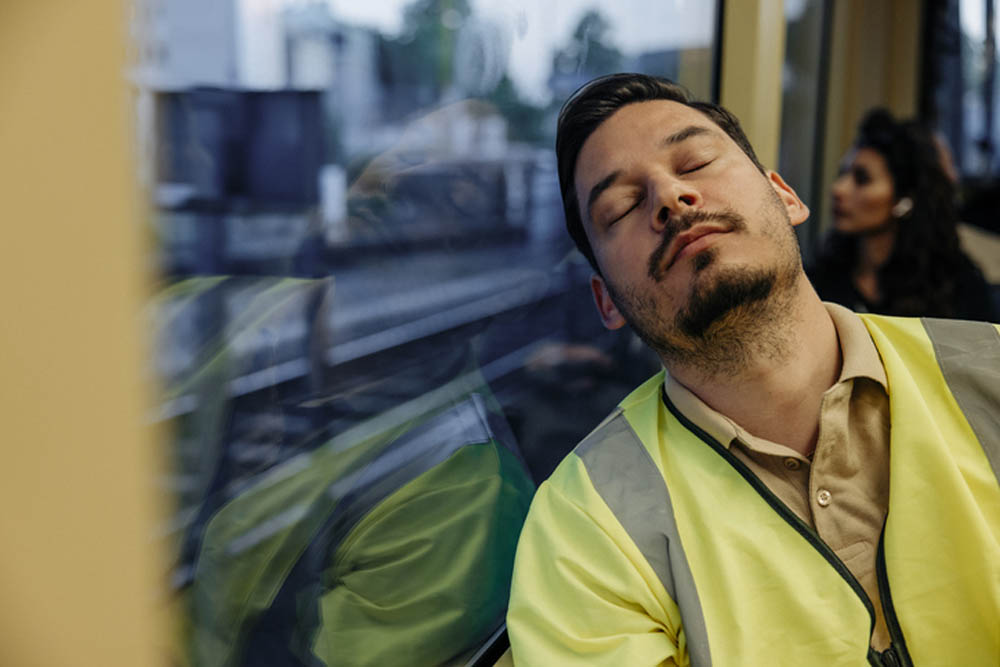 Man wearing a high-visibility yellow vest sleeping against the window of a moving train, with his reflection visible in the glass and a blurred cityscape outside.