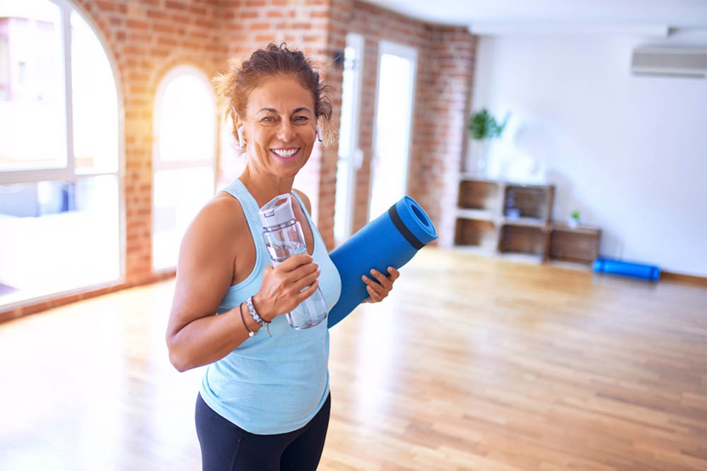 Smiling woman holding a rolled-up yoga mat and a glass of water, standing in a bright studio with wooden floors, brick walls, and large windows.