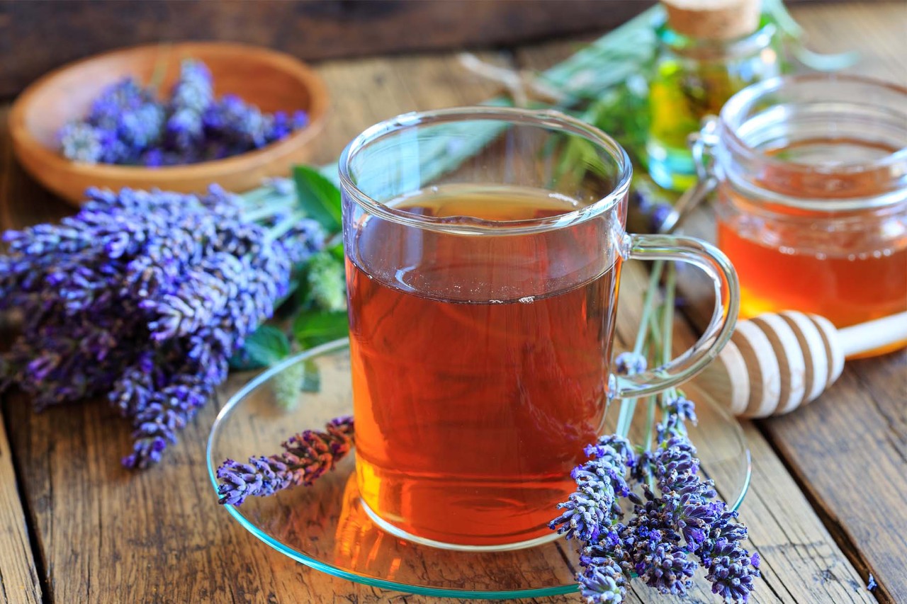 A glass of lavender tea on a plate, surrounded by fresh lavender sprigs and jars of honey, set on a rustic wooden table.
