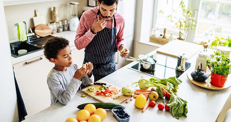 Father and young son preparing a healthy meal together in a bright kitchen, surrounded by fresh vegetables and fruits on the counter.