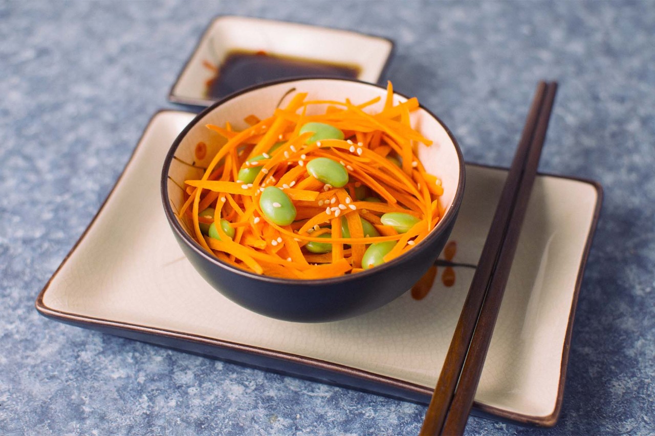 A bowl of shredded carrot salad mixed with edamame, drizzled with sauce, placed on a decorative plate alongside a small dish of sauce and chopsticks, all set against a blue background.