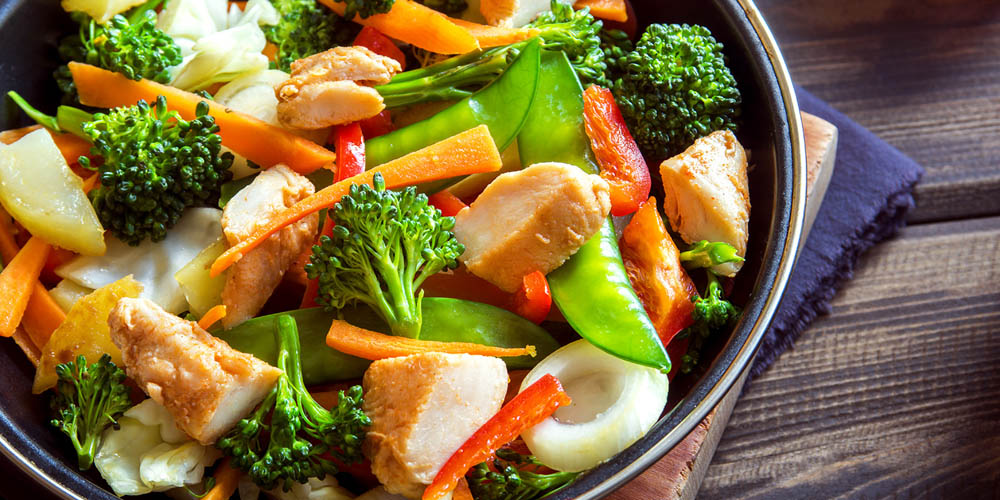 A bowl of stir-fried vegetables including broccoli, carrots, and snap peas, with chunks of chicken, arranged on a wooden surface.