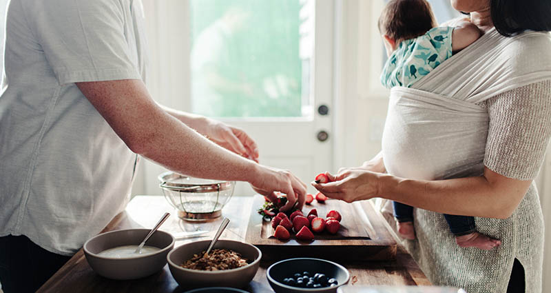 A caregiver holding a baby in a wrap while preparing food at a kitchen counter, with bowls of ingredients and fresh produce visible, as they engage in a collaborative cooking activity.